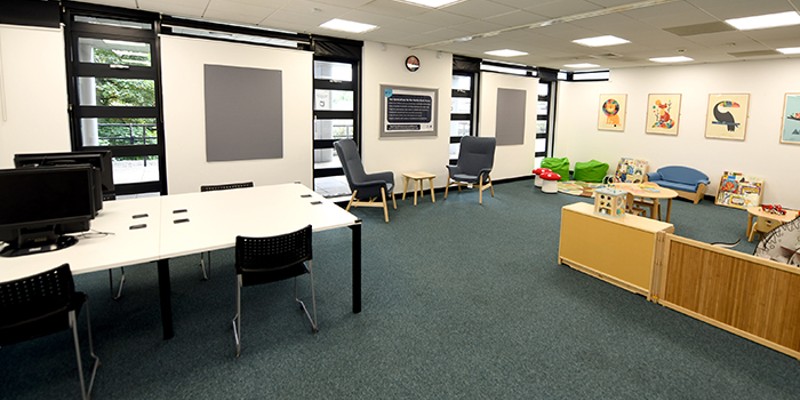 A view of the family study room, showing four desk spaces. Two of the desks hold desktop computers. There are also two armchairs at a coffee table and the children's area in the background with bookcases, toys and beanbags.